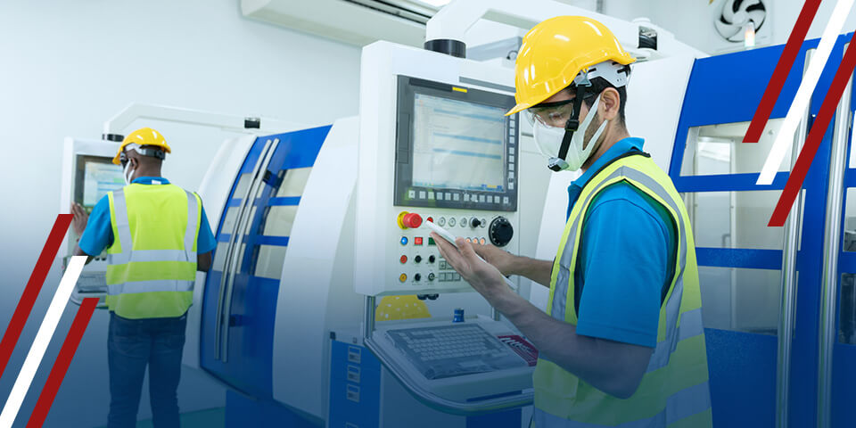 A worker operates a machine's control panel in a factory. He wears safety gear; another worker is visible in the background.