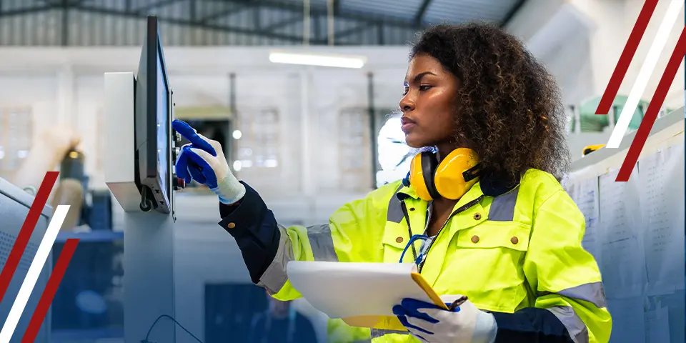 A woman in a lime-green safety jacket operates a digital screen in an industrial setting. She wears gloves and earmuffs, holding a clipboard.