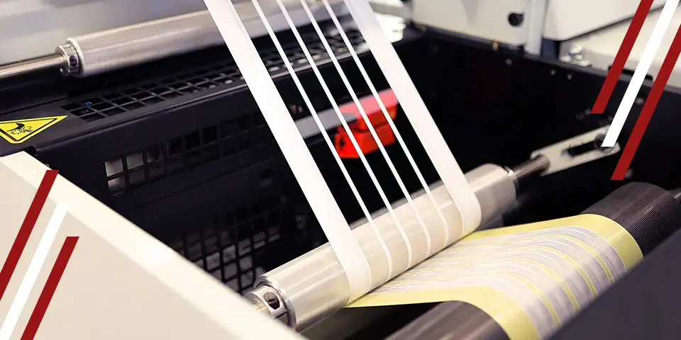 A label printing machine processes continuous label stock. Rolls of labels feed through rollers, creating a continuous stream of printed labels. The machine is part of a larger industrial process.