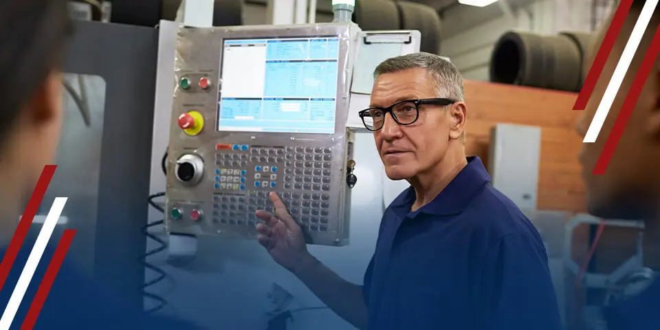 A man operates a machine's control panel, gesturing to explain its functions, in a factory setting.